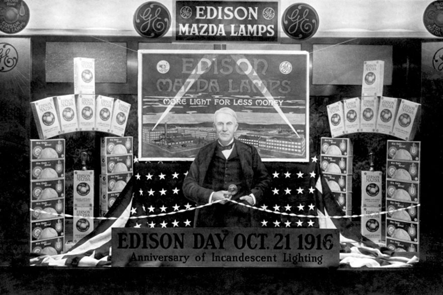 Ασπρόμαυρη φωτογραφία ενός άνδρα που στέκεται σε ένα πάγκο με λάμπες που γράφει Edison Mazda Lamps